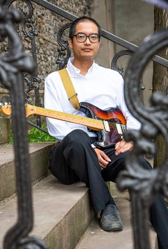 Ken Luk sitting on steps holding an electric guitar.